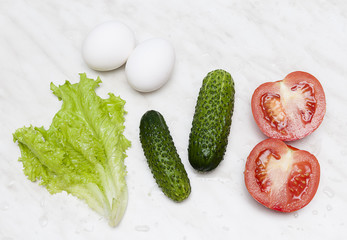 Fresh vegetables - tomato, cucumber, leaf salad - and eggs are ready for salad on light marble background 
