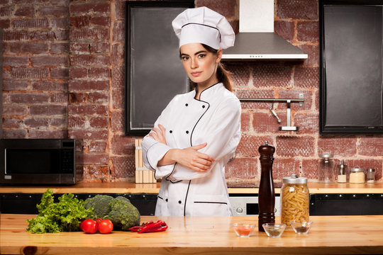 Female chef cook standing in the kitchen near wooden table with pasta, salad, broccoli, tomatoes, pepper, pepper mill and spices
