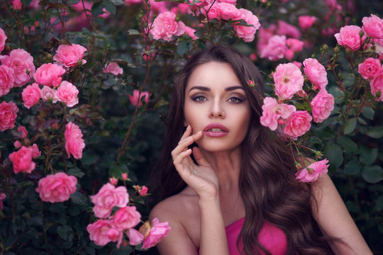 Fashion style beauty romantic portrait of young pretty beautiful woman with long curly hair posing between pink roses. Stunning girl looking at you or in camera