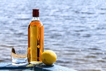 Bottle whiskey and tumbler with lemon on  shore of lake