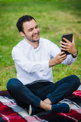 Modern young hispanic man taking a selfie at the park, with his tablet.