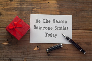 Be The Reason Someone Smiles Today  written on paper with pen,red gift box and wooden background...
