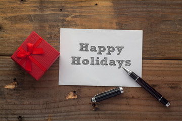 Happy Holidays written on paper with pen,red gift box and wooden background desk.