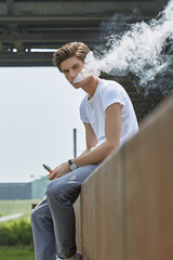Junger Mann in T-Shirt und Jeans sitzt e-Zigarette rauchend auf einer rostigen Absperrung unter...