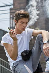 Junger Mann in T-Shirt und Jeans sitzt e-Zigarette rauchend an einer Bahnstrecke und bläst lässig eine weiße Dampfwolke. - obrazy, fototapety, plakaty