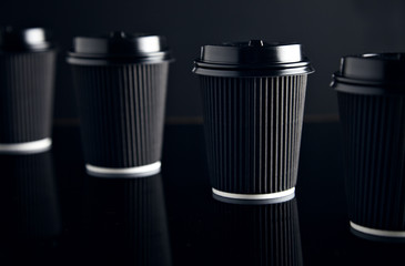Closeup four black take away cardboard paper cups closed with caps isolated on black and mirrored. Retail mockup presentation