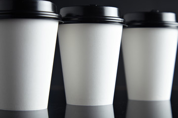 One focused paper cup for hot drinks between two unfocused presented on black back and mirrored, closed with caps, close focus mockup