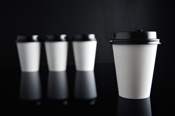 One focused white coffee take away paper cup ahead others unfocused, all closed with black caps presented on black and mirrored. Retail mockup presentation