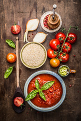 Dry Quinoa in ceramic bowl with tomatoes sauce and fresh ingredients for cooking, top view.  Quinoa preparation on rustic wooden background.