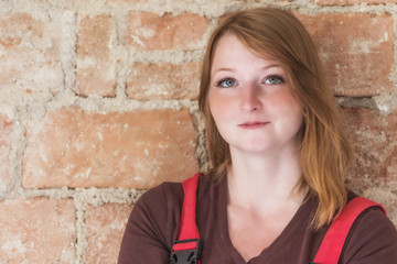 Portrait of smiling redhead young woman dressed in red overall is standing  in front of an old brick wall. Woman is looking at the camera.