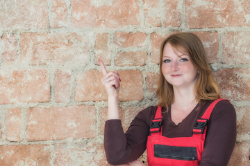 Attractive young redhead woman dressed in red overall is standing  in front of an old brick wall. Woman is looking at the camera and is pointing the finger at the wall.  