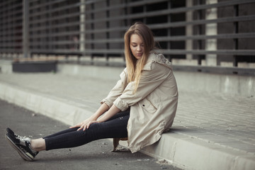 красивая девушка и бежевом плаще и джинсах сидит на тротуаре