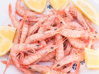 A plate of pink cooked shrimps. They are in their shells and their black eyes and feelers can be seen. The plate is on a green table outside.