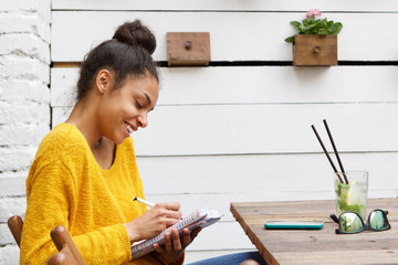 Happy young african woman sitting at cafe and writing notes