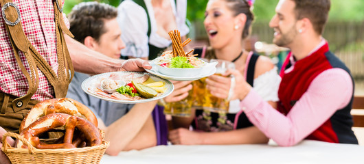 Kellner serviert Essen in bayerischem Biergarten, Freunde essen und trinken im Hintergrund