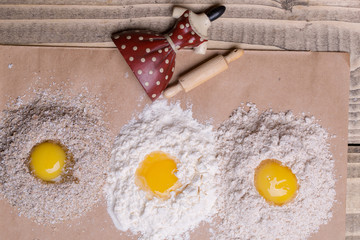 Three eggs broken on heaps of white and whole wheat flour bran with red dress porcelain figurine toy culinary utensils rolling pin on timber background