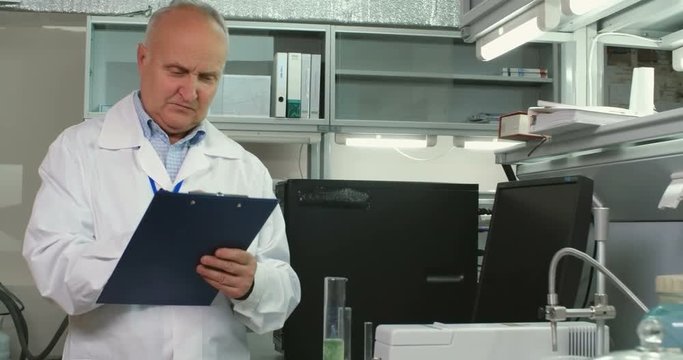 Senior chemist standing in laboratory, looking at two different chemical substances in glass tubes and writing down description of them