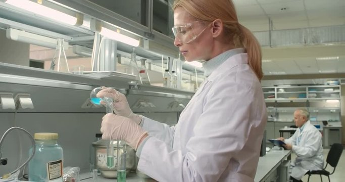 Female scientist pouring two chemical substances in one tube while senior chemist sitting behind her and writing something