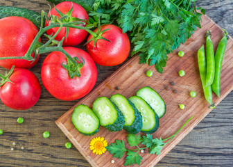 Cluster of ripe red tomatoes with water drops, sliced cucumber on wooden cutting board, bunch of parsley, green peas and peppercorn on the table, fresh vegetables, salad ingredients