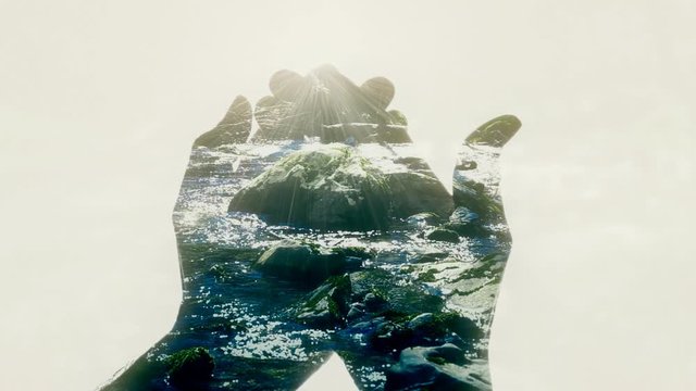 A pair of crossed hands forming a cup appear and reveal a scene in double exposure: flowing water from a river in a forest, bright glares from the sunlight.