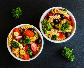 Salad: pasta fusilli, black and green olives, cherry tomatoes, red onion and parsley. Dressing: olive oil and lemon juice. In white bowl. Black stone background. Top view.