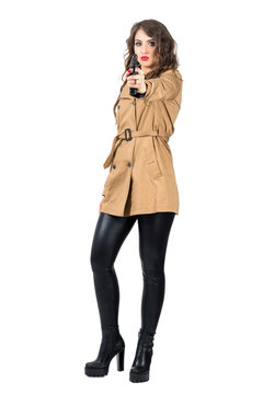 Sexy dangerous wavy hair spy wearing coat aiming weapon at camera. Full body length portrait isolated over white studio background