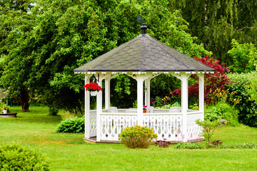 Lovely white gazebo with red flowers hanging from a basket. Trees and shrubs in the background. Fine garden with green grass. - 114432516
