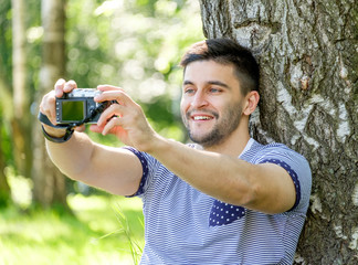 Portrait of young hipster man with camera outdoors. Young male photographer making selfie on summer day.