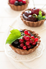 Close up of chocolate tartlets with chocolate cream, fresh strawberries, raspberries, blueberries, red currants and cherries on white wooden background. Selective focus