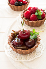 Close up of chocolate tartlets with chocolate cream, fresh strawberries, raspberries, blueberries, red currants and cherries on white wooden background. Selective focus