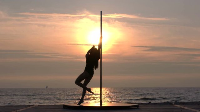 Silhouette of graceful girl dancer during fitness pole dance on the beach in the summer evening at sunset flexible poledancer performing advanced pole tricks and spins against the setting sun part_S2