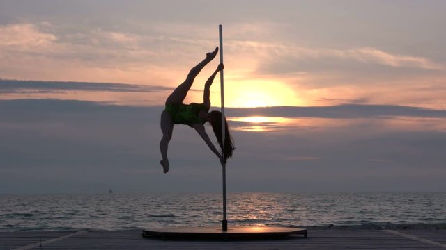 Strong girl performs fitness pole dance on the beach at sunset in the summer evening flexible poledancer against the glowing sun in the sky