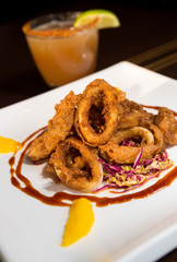 Beautiful golden crispy deep fried calamari rings, served on a white plate, with orange segments and a cocktail out of focus in the background.