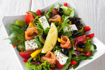 Mediterranean salad with salmon and brie top view. Appetizing salad with salmon, brie, olives and vegetables, served with lemon slice in white bowl on wooden table.