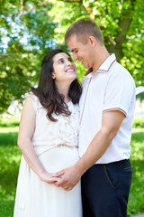 Fototapeta premium pregnant woman with husband posing in the city park, family portrait, summer season, green grass and trees