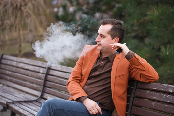 A young man sits on a bench and smokes an electronic cigarette.