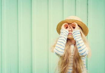 Cute little girl stands near a turquoise wall in boater hat and  looks invented binoculars. Space...