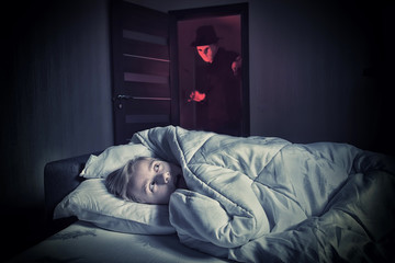 Nightmare. Scared boy lying in the bed while the masked stranger standing in a doorway