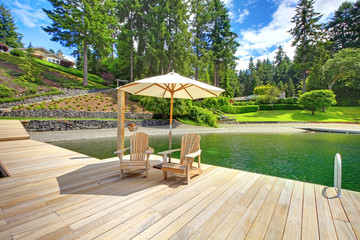 Two adirondack wooden chairs with umbrella on dock facing beautiful landscape.
