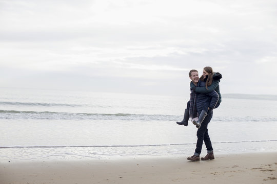 Young Man Giving Woman Piggyback Ride Along Beach