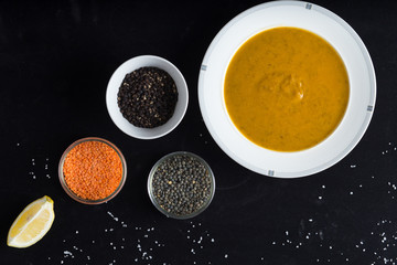 Cream soup of assorted lentil. Yellow and green lens, spices as raw for meal and lemon on black background. Healthy, appetizing, delicious, vegetarian food. Top view, copy space.