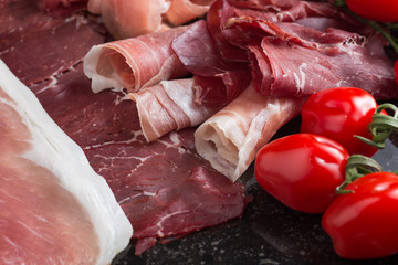Jambon mix. Ham. Traditional Italian and Spanish salting, smoking, dry-cured dish - jamon Serrano and prosciutto crudo sliced with herbs and tomatos on dark stone background. Copy space. Closeup. 
