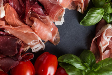 Jambon mix. Ham. Traditional Italian and Spanish salting, smoking, dry-cured dish - jamon Serrano and prosciutto crudo sliced with herbs and tomatos on dark stone background. Copy space. Closeup. 
