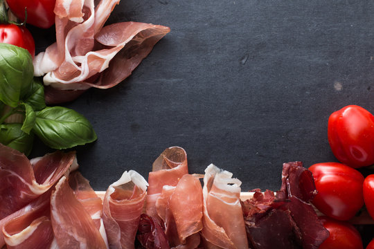 Jambon mix. Ham. Traditional Italian and Spanish salting, smoking, dry-cured dish - jamon Serrano and prosciutto crudo sliced with herbs and tomatos on dark stone background. Copy space. Closeup. 
