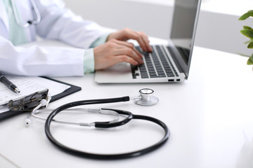 Close-up of stethoscope is lying on the table near female doctor typing on laptop computer