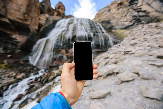 Male hand with a smartphone on the background of mountain waterfall
