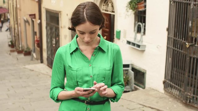 Attractive Young Brunette Woman in Light Summer Dress on Streets of European Town. She is looking at her Mobile Phone and using it.