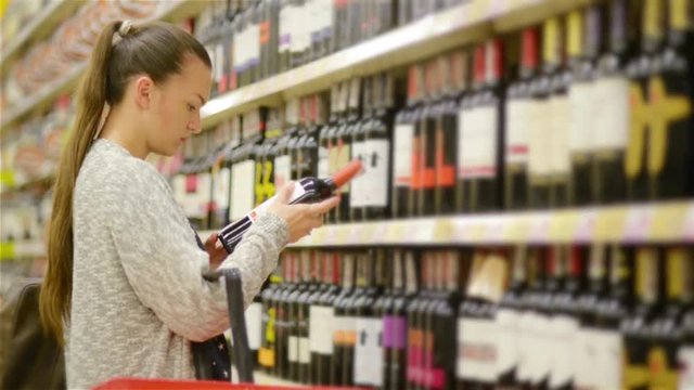 Woman shopping for wine or other alcohol in a bottle store standing in front of shelves full of bottles with a serious expression as she tries to make up her mind