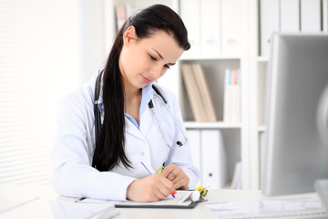 Young brunette female doctor sitting  with clipboard near window in  hospital and filling up medical history form.