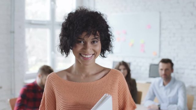 Closeup of happy young woman looking at camera standing with documents in office
